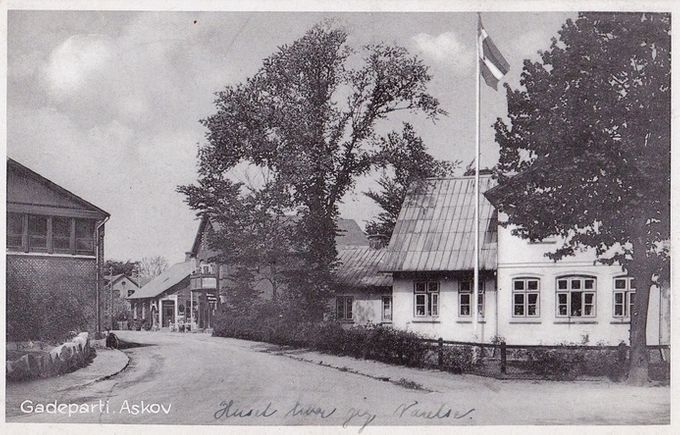 Nummer tre hus fra højre er Askov Boghandel. Det var utrolig dejligt at komme derind. Der var bøger på hylder og stabler af bøger på gulvet. Boghandleren viste lige præcis hvor en titel på en bog skulle findes. Ellers fik man lov til selv at lede. Huset med flaget, var en kafferesturant, som jeg var inde flere gange,for at få en kande kaffe. Huset ved siden af Boghandleren, havde en frisør i mange år. Nu har jeg hørt at det er blevet nedrevet.