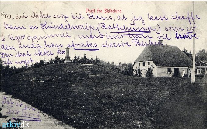 Poststempel 1907.
Parti fra Skibelund.
Der står på postkortet.
Vil du ikke sige til Hans, at jeg kan skaffe ham en Hundehvalp (Rottehund.)  Som er 6 Uger gammel men hvis han vil købe den, må han straks skrive Besked. Den skal ikke koste noget Det er en Fox Terri.
Ja, så er den besked givet videre fra 1907.
Det er det første billede jeg ser hvor der er mennesker ved det lille hus. Mon ikke det var   restauration dengang.
Og højen til venstre for det lille hus har benævnelsen kæmpehøjen.
Stenen på højen er den første sten i Skibelund. Det første monument, der blev opstillet i Skibelund Krat, står til minde om J.C. Lindberg (1797-1857).  Jacob Christian Lindberg (14. januar 1797 i Ribe – 10. december 1857) var en dansk præst og politiker. Symbolsk står det på en gravhøj fra bronzealderen.
Der står.
De begravede i gravhøjene.
Menneskene i ældre bronzealder satte gang i et storstilet højbyggeri. I gravhøjene blev der værnet om den døde person, både ved graven selv og ved den måde, hvorpå man gjorde højen synlig. Højene sikrede samtidig erindringen om de døde i kommende generationer. Igennem hele bronzealderen blev højene brugt igen og igen til begravelser. Den enkelte høj eller gruppe af høje kom til at udgøre en gravplads, som man i århundreder vendte tilbage til.