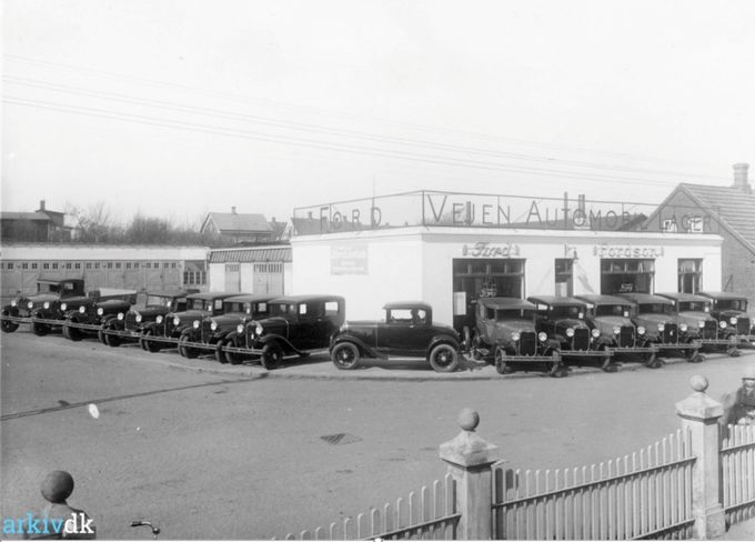 Vejen Automobil Lager på hjørnet af Askovvej og Søndergade. 
Årstal 1929.
Fotograf K. H. Kock, Vejen. 
......
Et sprit nyt billede fra starten af Ford i 1925 ved krydset Søndergade og Askovvej.
Fotografiet er taget inde fra  Ole  Haagensens villa på hjørnet af Søndergade, Plantagevej,   hvor der i gamle dage var et hvidt stakit rundt om den store have, der blev mindre da Plantagevej blev udvidet.
Haagensens villa var født hvid, men har haft flere farver. Gul bl.a.
Jeg ville gerne have lidt kulør på billedet  men det gik ikke, halvdelen af bilerne blev røde. Og jeg kan huske at i de første mange år var alle bilerne HGF sorte. Så da den første røde bil kom til Danmark blev der rigtig kigget på den. I dag er hele  farvespektret repræsenteret i bilparken.
Oppe bagved til venstre ligger som det første hus i Allegade/Askovvej  Karetmager Petersen,  han er i den grad blevet glemt. Hans kone var umådelig flink at tale med. Jeg mener vi kaldte ham Juler Pedersen, nok et øgenavn på grund af at han lavede hjul, også til Ford, hvor han lavede træarbejde til anhængere. Han var fodsboldspiller på førsteholdet i 1930erne, i Vejen. 
Og det andet hus til højre er mine bedsteforældres hus nummer 17.
Det var det første hus i Vejen, jeg kom i da jeg var meget lille.
Jeg er født i 1946, så det er lidt sjovt at Allegade 17. er fra 1929.
Huset til højre for mine bedsteforældres hus var hvid. Der boede familien Terp. For et par uger siden blev det nedrevet. Og det var for en gang skyld en god beslutning.
Jens Aksel Kjær skriver.
Dejlig at se Karetmager Petersens hus. Udover at være karetmager røgede han også kød for folk. Jeg har så gode minder fra dette når vi fik røget en skinke og bacon hos ham før jul. Det var en 