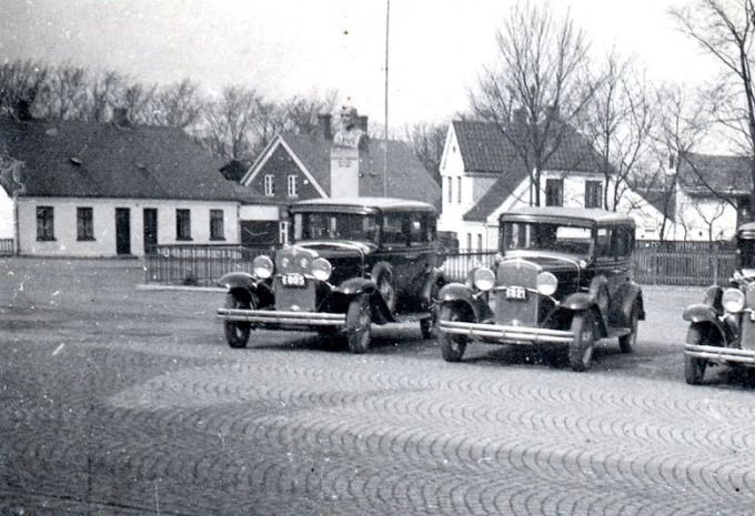 Et dejlig billede af en tid der var.
De første taxier ved Banegårdspladsen.
Der står ikke hvilket årstal det er, men de høje HGF`er   var de første biler man så i gadebilledet omkring 1929. To der var med fra starten var Jens Skov og Hans Olander.
Det må have været et stort arbejde at lægge brosten i hele byen.  Brostenene i Vejen kom i 1924.
Visit Vejen skriver.
 Foran Vejen Station knejser byens 