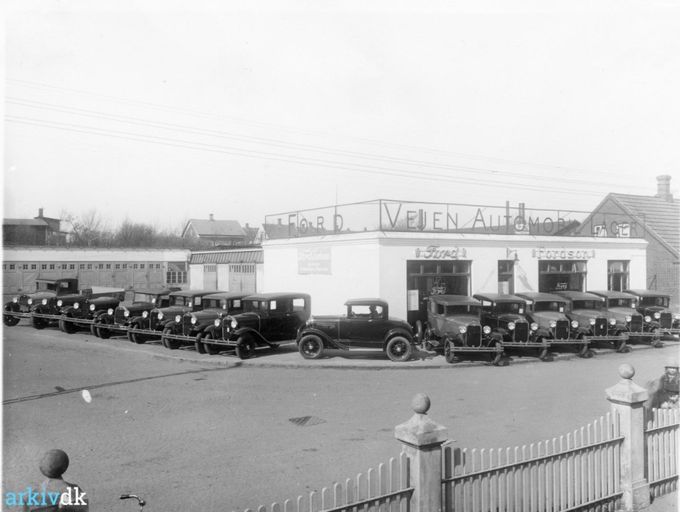 Vejen Automobil Lager på hjørnet af Askovvej og Søndergade. 
Årstal 1929.
Fotograf K. H. Kock, Vejen. 
......
Et sprit nyt billede fra starten af Ford i 1925 ved krydset Søndergade og Askovvej.
Fotografiet er taget inde fra  Ole  Haagensens villa på hjørnet af Søndergade, Plantagevej,   hvor der i gamle dage var et hvidt stakit rundt om den store have, der blev mindre da Plantagevej blev udvidet.
Haagensens villa var født hvid, men har haft flere farver. Gul bl.a.
Jeg ville gerne have lidt kulør på billedet  men det gik ikke, halvdelen af bilerne blev røde. Og jeg kan huske at i de første mange år var alle bilerne HGF sorte. Så da den første røde bil kom til Danmark blev der rigtig kigget på den. I dag er hele  farvespektret repræsenteret i bilparken.
Oppe bagved til venstre ligger som det første hus i Allegade/Askovvej  Karetmager Petersen,  han er i den grad blevet glemt. Hans kone var umådelig flink at tale med. Jeg mener vi kaldte ham Juler Pedersen, nok et øgenavn på grund af at han lavede hjul, også til Ford, hvor han lavede træarbejde til anhængere. Han var fodsboldspiller på førsteholdet i 1930erne, i Vejen. 
Og det andet hus til højre er mine bedsteforældres hus nummer 17.
Det var det første hus i Vejen, jeg kom i da jeg var meget lille.
Jeg er født i 1946, så det er lidt sjovt at Allegade 17. er fra 1929.
Huset til højre for mine bedsteforældres hus var hvid. Der boede familien Terp. For et par uger siden blev det nedrevet. Og det var for en gang skyld en god beslutning.
Jens Aksel Kjær skriver.
Dejlig at se Karetmager Petersens hus. Udover at være karetmager røgede han også kød for folk. Jeg har så gode minder fra dette når vi fik røget en skinke og bacon hos ham før jul. Det var en 