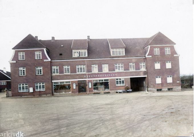 Vejen Brugsforening, Banegårdspladsen 3, Vejen. Bygningen er opført i 1913.
Den var nu flot den gamle Brugsforening på Banegårdspladsen.
Her med lidt farve.
Til højre i brugsforeningens vindue fylder den store kaffemaskine godt i vinduet.
Til venstre på bygningen kan jeg huske at der var en flot vin, der om efteråret stod med røde farver.
Jeg tror at billedet her er før 1946.
Der var rigtig god plads på Banegårdspladsen.
Det var nogle tunge træ trillebøre man købte den gang. Der må jeg sige at de letvægt trillebøre  man køber i dag er en god ting.
Jeg kan ikke huske indkørslen ind til højre.
I dag er der ikke sådan en ro på som den gang.
Til højre var det til VesterAlle og Jernbanegade.
Og til venstre var det de sorte  træhusene der hørte til Togstationen.