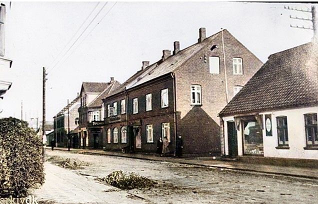 Endnu et spændende billede fra Vejen arkiv.dk
Det er et meget gammelt billede af Søndergade. Det var før der blev lagt brosten i Vejen by. I 1924 brolagde man gaderne i Vejen.
Den første butik 1890 til Ebbe Lundgaard startede ved Søndergades Viktualie-, Paalægs- og Delikatesseforretning ved K. Lundgård siden ved Katrine Marie Lundgaard 1913.
Hun havde butikken frem til 1957.
Siden kommer Forretningsoverdragelse.
Det meddeles herved, at
K. Lundgaards paalægs- og viktualieforretning, Søndergade 17, Vejen,
fra og med mandag den 30. ds. overdrages til slagter Peter Knudsen.
Han blev svend og senere mestersvend
hos slagtermester Karl Knudsen Søndergade 6. Han døde alt for tidlig i 1982, 55 år gammel.
Dobbelthuset til venstre må være Bagerforretningen og Palmehaven på hjørnet af Fredensgade.
De to næste huse på den anden side af Fredensgade tør jeg ikke kloge mig på. Jeg mener at Tøjbutikken ved Jensen og Petersen kom senere i 1940.
Det sidste store hus med flagstangen til venstre mener jeg må være det nybyggede Posthus i 1861.
Det er lidt sjovt at se hovedgaden med jord..
Jette Asmussen.
Jeg husker fru Lundgaard, fra da jeg var barn. Hun var vores nabo indtil hun flyttede på plejehjem. Vi boede i nr 15. Det var i midt tresserne.
Nu bor jeg ved siden af hendes barnebarn Else Lundgaard ( 95 år ) i Rødovre. Verden er lille.