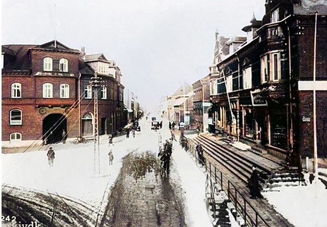 Nørregade i Vejen, fotograferet fra viadukten mod nord, omk. 1940.
Et nyt farvet vinterbillede, hvor Laurits Andersen, Byens Hjørne, til venstre ikke har fået forretningen endnu ved Hansens Hotel. Til gengæld er der porten ind til Hansens Hotel. Porten var høj og vid så hestekøretøjer kunne køre ind i hotellets stald hvor der i ældre tid var dyreauktioner derinde.
De 2 første vinduer til venstre fra porten var der en krostue, til dem der kom på auktionerne. Der var fire trin op til krostuen.
I 1940erne var der gennemgang ved porten, så man kunne køre lige igennem i bil.
Porten var høj og vid så hestekøretøjer kunne køre ind i hotellets stald.
Det er sikkert på den tid hvor de første biler begyndte at køre i Vejen by.
Til højre er det de 5 trappetrin op til Fotograf K. H. Koch og helt til højre ved gavlen var det Telefoncentralen. Til venstre var det Vejen Materialhandel. Forretningen, som grundlagdes 1885, overtoges i 1939 af Aage Nielsen. Jeg læser at i starten var det Hotelejer H. C. Hansen 1858-1924 på Hansens hotel der ejede bygningen.
Ja det var glat der om vinteren når man cyklede ned under viadukten. Jeg har ligget med knuste æg der som jeg havde købt hos æg pakkeriet i Ahornsgade. Dengang var der noget der hed knækæg, som bagerne kunne købe meget billigt.
På den anden side af Ahornsgade var den flotte bank.
Til højre nede ved vejen er der de sjove trappetrin op til fortovet. Jeg har ikke fundet ud af hvorfor det var der, men tænker at man gik under tunellen inden der kom biler i Vejen.
I 1940erne var det de meget kolde snevintre der var der.
31,2 minusgrader er Danmarks officielle kulderekord.
Bertha Kirstine Christiansen.
Der var en dør ind mellem Vejen Materialhandel og urmager Schmidt. Indenfor døren trappe til 1.sal, hvor tandlæge Sv.O.Nyvad havde klinik. Jeg blev uddannet der som klinikassistent 1955-1958. Fru Ingemann Jensen var også tandlæge der.