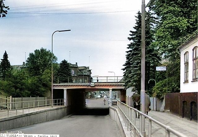 Et farvet billede af Viadukten, inden huset til højre blev nedrevet  i 1974, hvor Bogtrykker Herluf Frandsen boede i  Søndergade nr. 1, Vejen. 
  Huset lå på hjørnet af Søndergade/Mindevej. Fotograferet omkring 1970.
Der står.
Det startede med Hans W. Jäger. Vogn & Beslagsmed ca. 1938.
Og ca. i 1950 var det Vejen Smédie- & Maskinlager, ved Ole G. Madsen.  
Da jeg begyndte at komme i Vejen, var det  H. P. Frandsen, der havde Bogtrykkeri og beboelse der. Og der var stensætningen væk, med planter, og den sorte mur i stedet.  H. P. Frandsen, startede i Vejen 5. marts 1959 med Vejen Bogtrykkeri. Da var jeg 12 år. Pludselig skulle huset nedrives, og  H. P. Frandsen, måtte i gang med at finde et andet sted til sit trykkeri. 
Det var dengang der stadig var ro på i hovedgaden.
