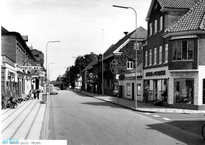 Et utroligt dejlig billede af Søndergade.
Til højre starter det med Fredensgade, og vores tøjforretning fra 1946 og lang tid op i 1960erne.  Jensen & Petersen's Herreekvipering. Så kommer først Elektrikeren ved Orla Jensen i 1966 og Frede Slot som tager over i 1979.
Så kommer Posthuset med den røde postkasse. Dens tids Messenger, hvor man kunne nå at fortryde et rapt svar. Med en flot efeu der var kendetegnende for Vejen i den tid.
Helt tilbage i 1938 var det ved siden af Posthuset Skomager Marinus P. Jensen og Frk. Anna Jensen.
til 1957.
Så er det Antoni  Boysen, Kronhøjs efterfølger, og siden sønnen Hans Boysen.
Inden Mindevej er det Bertha Christensens Trikotageforretning i Søndergade nr. 1.
Til venstre er det  Henry Andersen,  Isenkram Glas Porcelæn 1954, hvor vi også handlede.
Før da var det i  1938 Isenkræmmer Chr. And. Hübbe.
Så kommer Juel Pedersen, Konfektion Kjoler og Sengeudstyr 1954. Da Juel Pedersen og hans kone stoppede med forretningen,  blev han lærer på Handelsskolen.
Den sidste butik inden Jyllandsgade er Karl Egon Haagensens Købmandsbutik som vi handlede meget ved.
Det  var en dejlig og rolig tid i 1950erne end i dag hvor det nærmest er et ragnarok at færdes i Vejen.