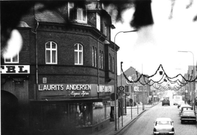 Til højre op af de 5 trappetrin kan man lige se
Vejen Materialhandel ved ved Aage Nielsen.
Forretningen startede i 1885. Vi var tit hos ham hvor jeg blandt andet købte kæmpe duftende hirsekolber til mine undulater, som han hentede i kælderen. Og fik dem udleveret i brune papirsposer. Ja der var der ikke noget der hed plasticposer dengang i starten af 1950erne.
Så kommer Ahornsgade, og nr. 8 Banken for Vejen og Omegn.
Og ved siden af er det Bruun- Møllers Boghandel. Ja man ser ikke ure mere i bybilledet ej heller telefonbokse som man havde meget brug for den gang. Desværre var der meget hærværk i telefonboksene.
I 1974 skulle Nørregade nr. 10 Bruun-Møllers boghandel nedrives. Desværre røg muren også til Vejen Bank med i købet. Så efter nogle grundige overvejelser rev man også den flotte bygning ned, nu man alligevel var godt i gang.
Til venstre ved Hansens Hotel er det Manufakturhandler Laurits Andersens forretning. Og her er sikkert starten på nedrivningerne i Nørregade. Da man er kommet til Bager Sørensens butik der dog stadig står der endnu.
Hvorfor man ikke stoppede nedrivningerne her er mig en gåde, som jeg sikkert aldrig får svar på.
Selv som gammel Vejen borger kan jeg ikke finde ud af hvordan det nu var hvis jeg går dernede her i 2022. Så hvis der kommer nye mennesker til Vejen har de ikke en kinamands chance for at vide hvordan det nu var.
Dorthe Holm Jensen.
Sådan husker jeg Vejen, og guirlanderne ved Juletid. Det er synd at der ikke var nogen medlemmer i Vejen Kommune som sad i planlægning af Vejen’s udvikling som kunne have holdt lidt igen med nedrivning på det tidspunkt.
