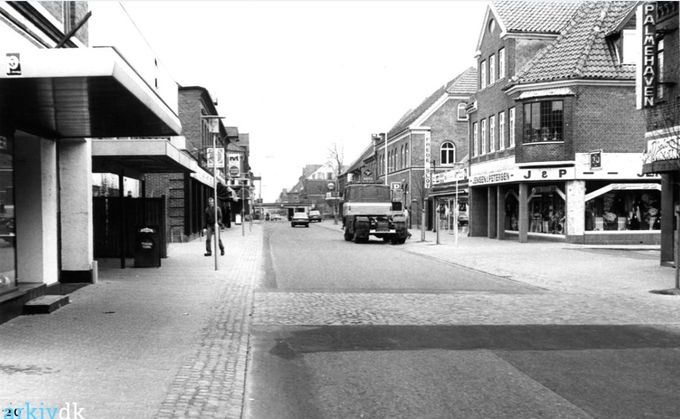 Et nyt flot billede af Søndergade, med Palmehaven ved restauratør N. J. J. Varming helt til højre, og senere A. og J. Georgsen. Søndergade 13.
Så kommer Fredensgade og Jensen & Petersen's Herreekvipering, som vi brugte fra 1946 og fremefter. Søndergade nr. 7 er opført i år 1872.
Så kommer Elektrikeren ved Orla Jensen i 1966, og senere overtog El- Installatør Frede Slot butikken omkring 1979.
Så kommer Posthuset, hvor man kunne fortryde hurtige svar inden man havde skrevet et brev og fået det lagt i Posthusets Postkasse.
I dag klikker man på en tast i sin computer, og lynhurtigt er et god eller dårlig svar ude..
I 1986 flyttede Vejen Posthus til ny bygning i Jyllandsgade nr. 6.
Og så kommer Erna Andersens Vejen Foto. Hun og hendes mand startede ved viadukten i Nørregade 4. efter Urmager og guldsmed Arne Schmidt, hvor Fotograf P. H. Koch først havde sit domicil. Når man skulle op og fotograferes skulle man op af en temmelig mørk trappe der duftede af Fotografens fremkaldervæsker.
Til venstre med bygningen med det store udhæng kan jeg ikke huske.
Men efter det er Det Tove og Volmer Jensens dyrebutik hvor man kunne købe alt fra hånd tamme undulater til akvariefisk.
Så kommer efter Isenkræmmer Henry Andersen og senere Schmidt's Isenkram i Søndergade 10.
