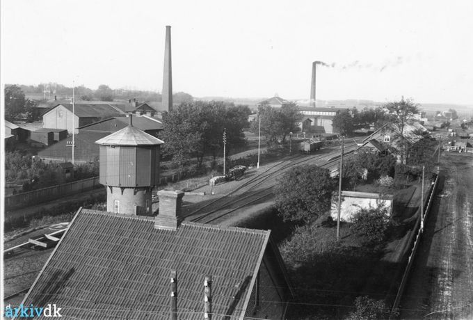 Baneterrænet, fotograferet fra Højskolehjemmets tag, 1916
Årstal 1916
Fotograf K. H. Kock, Vejen
,,,,,,,,,,,
Et nyt flot billede fra den gamle Banegård.
Helt til højre er det den endnu  jordbelagte vej op til  Vejen banegård, hvor Hansens hotel ligger overfor.
Jeg er sikker på at huset forneden i billedet må være Ledvogterhuset nr 7.  som blev fjernet samme år som billedet her blev taget, da viadukten blev udgravet i 1916-17. Og som Hotelejer H.C. Hansen købte og fik opført igen i  Ahorngade nr. 5.
Hvor der står.
 Ahorngade 5, Vejen. Bygget af nedbrydningsmaterialer fra det gamle ledvogterhus ved jernbaneoverskæringen, hvor nu viadukten Nørregade/Søndergade er. Hotelejer Hansen købte huset og flyttede det. Nicolaj Pugh købte det i 1919 for ca. 5000 kr.
Og ved stationsbygningen var bygget et Vandtårn så  lokomotiverne kunne forsynes med vand.
Og til venstre for jernbanen ses forrest Cikorie fabrikken  Nørrejylland  og bagerst Margarine fabrikken  Alfa`s skorstene.
 Den gamle stationsbygning fra  1874 
blev nedrevet og i 1917 blev den flotte bygning man ser i dag opført, der blev  tegnet af DSB's arkitekt Heinrich Wenck. Der også havde tegnet flere togstationer, blandt andet Københavns Hovedbanegård fra 1911.