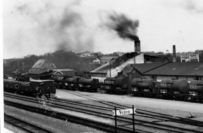 Tagpapfabrikken Phønix set fra banen i 1932.
,,,,,,,,,,,,,,,,,,,,,
Ja den sorte røg er fortid nu. Det døjer vi med i dag 2022.
Industrialiseringen startede i 1800 tallet. Det var med kul og kulværker der ikke var godt for
miljøet.
I 1932 var Vejen stadig omkrandset af landbruget.
Øverst midt i billedet kan man se det gamle vandtårn, der blev opført i 1923.
Og til venstre er det gården Baungaard. Og foran er det Kærdalsgaard. I dag Kærdalens Plejecenter.
Bag Phønix skorstene er det Skovvejen der førte op til Højbro der blev lavet i 1916.
Huset øverst til venstre med de to vinduer i gavlen er direktør Rudolf Frederiksens idylliske hus nummer 33, der er her endnu.
Direktør R. Frederiksen havde ikke langt til sit arbejde på Phønix.
Jørgen Schlünssen.
Det lille hus i forgrunden indeholdt en brovægt.
